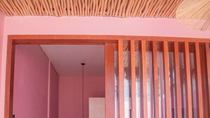 Una habitación rosa con techo de listones de madera, una puerta blanca y vistas a un baño con inodoro y lavamanos blancos.