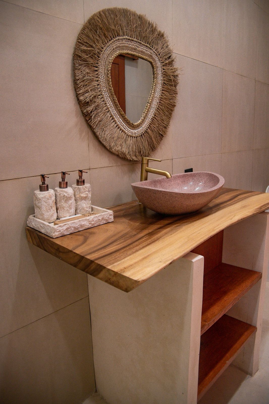 Een badkamer met een houten wastafelmeubel, een ronde spiegel met een rand, een roze stenen wastafel en drie zeepdispensers.