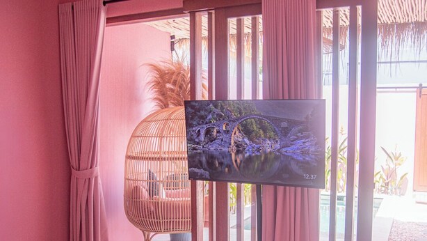 Habitación de hotel en tonos rosados con una cama, televisor y vistas a un área exterior tropical.