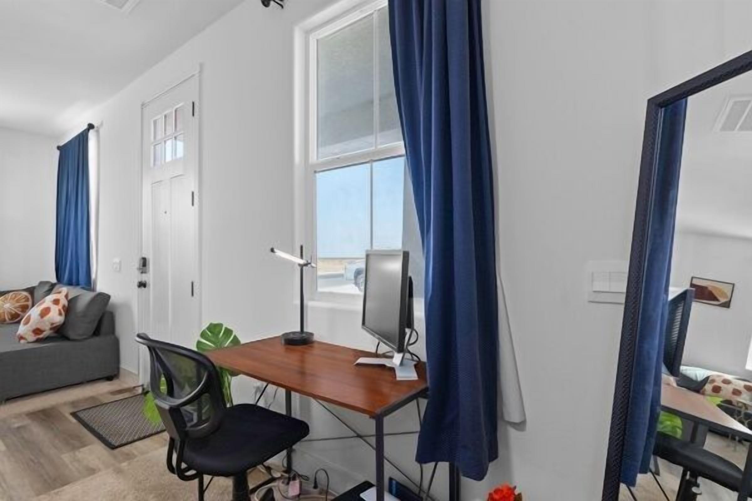 Una habitación moderna con escritorio, silla y ordenador. Hay una ventana con cortinas, un sofá con cojines y un espejo.