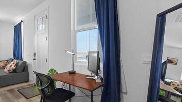 Una habitación moderna con escritorio, silla y ordenador. Hay una ventana con cortinas, un sofá con cojines y un espejo.