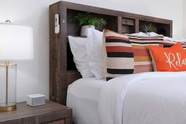 A neatly made bed with striped and solid pillows, a bedside lamp, and a small white speaker on a wooden nightstand.