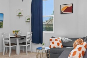 A modern living room with a dining area, a grey sofa with orange and white patterned cushions, a blue curtain, and a framed abstract painting on the wall.