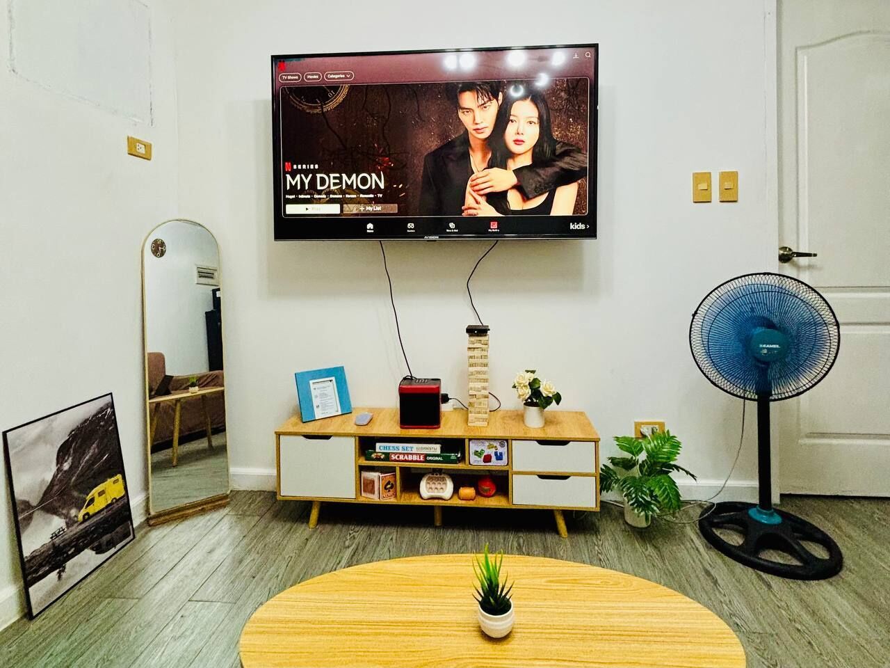 A living room with a TV mounted on the wall, a wooden cabinet with various items, a small table with a plant, and a standing fan.