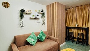 A living room with a brown sofa, two green pillows, a wooden coffee table, a small potted plant, a wall clock, a shelf with plants, a power outlet, and a curtain.
