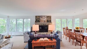 A spacious living room with a fireplace, large windows, a dining area with wooden chairs, and a patterned sofa.