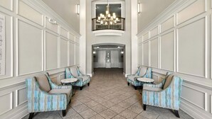Een hotel lobby met een hoge plafond, een kroonluchter en een centrale deur die naar een andere ruimte leidt. Er zijn twee zithoeken met gestreept meubilair en kleine tafels.