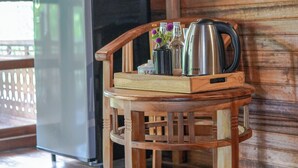 Una silla de madera con una bandeja que sostiene un hervidor, una botella y un florero con flores, ubicada en una cabaña con paredes de madera y un refrigerador al fondo.