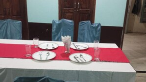 A dining table set with plates, cutlery, and glasses, covered by a red tablecloth, in a room with wooden doors and blue chairs. - Hotel Singhasan Haveli (Mandawa)