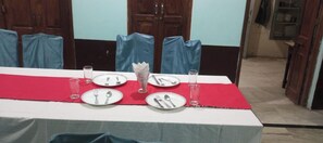 A dining table set with plates, cutlery, and glasses, covered by a red tablecloth, in a room with wooden doors and blue chairs. - Hotel Singhasan Haveli (Mandawa)