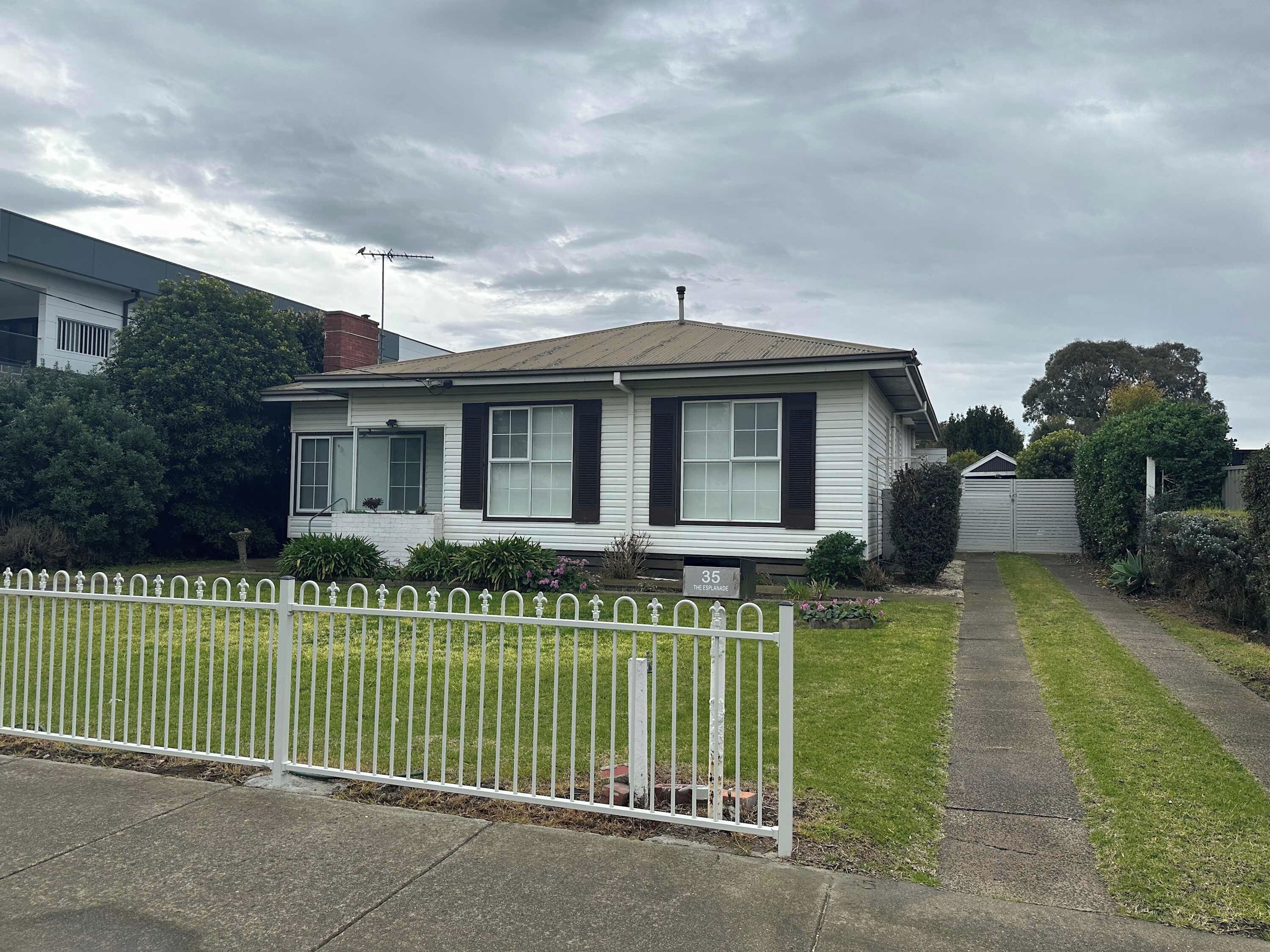 A single-story house with a white exterior and dark shutters, a front yard with a white fence, and a driveway leading to the garage.