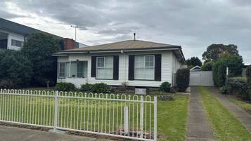 A single-story house with a white exterior and dark shutters, a front yard with a white fence, and a driveway leading to the garage.