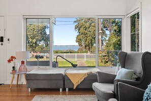 A modern living room with a grey sectional sofa, a small table with a lamp, and a view of a body of water through large sliding glass doors.