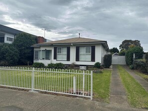 A single-story house with a white exterior and dark shutters, a front yard with a white fence, and a driveway leading to the garage.