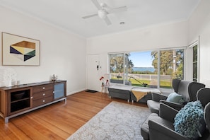 A modern living room with a wooden cabinet, grey sofa, and a large window offering a view of a garden and a body of water.