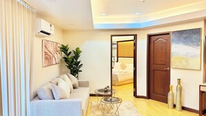 Una sala moderna con un sofá, una mesita de centro y una planta grande. Se ve una puerta que da a otra habitación con una cama.