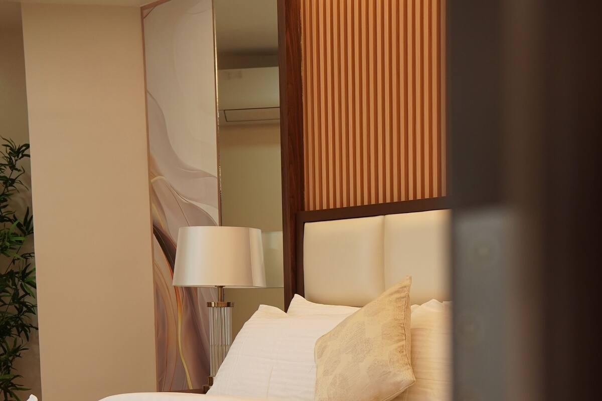 Un lit bien fait, avec du linge de lit blanc et une serviette pliée, une lampe de chevet et un miroir au mur.