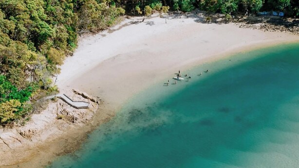 Eine Vogelperspektive zeigt einen Strand mit klarem türkisblauem Wasser und einem sandigen Ufer, auf dem sich einige Personen im Wasser befinden.