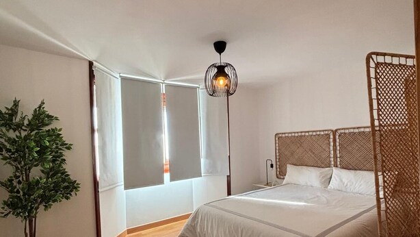 A bedroom with a large bed, a potted plant, a hanging light fixture, and a window with blinds. - Fuente del Tesoro Apartment (AN)