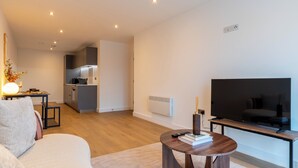 Una sala de estar moderna con un sofá, una mesa de centro, un televisor sobre un mueble bajo y una cocina al fondo.