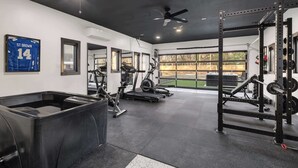 A gym with exercise equipment, a ceiling fan, and a large window with a view of the outdoors. - Renovated 7BR - Gym, Hot Tub, Sauna & Cold Plunge! (Southfield)