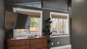 A room with a wooden dresser, a flat-screen TV, and two windows with white blinds. - Renovated 7BR - Gym, Hot Tub, Sauna & Cold Plunge! (Southfield)