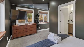 A bedroom with a bed, a dresser, a TV, and a window with a view of trees. - Renovated 7BR - Gym, Hot Tub, Sauna & Cold Plunge! (Southfield)