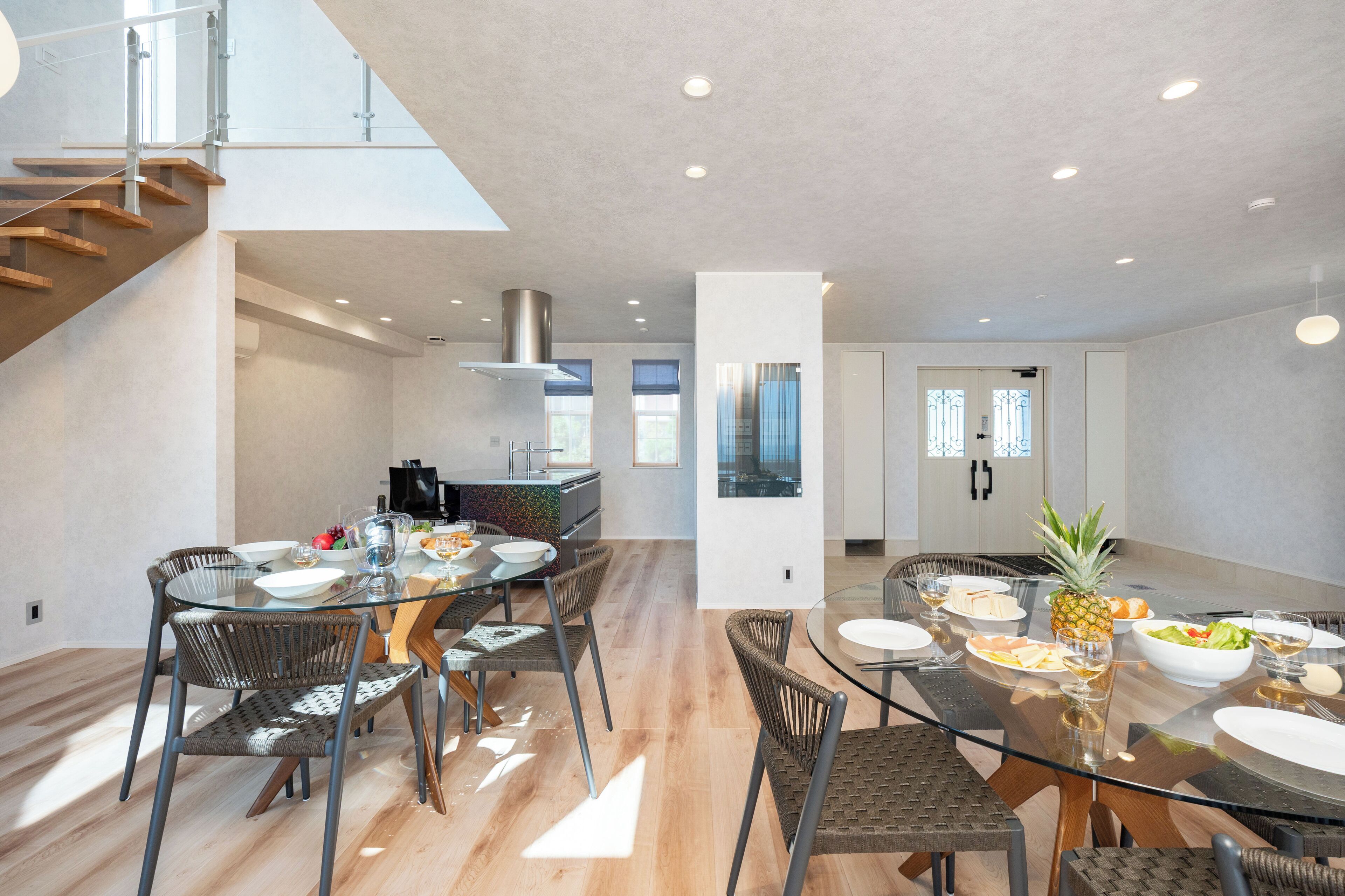 A modern kitchen with a dining area, featuring a glass table, wicker chairs, and a staircase.