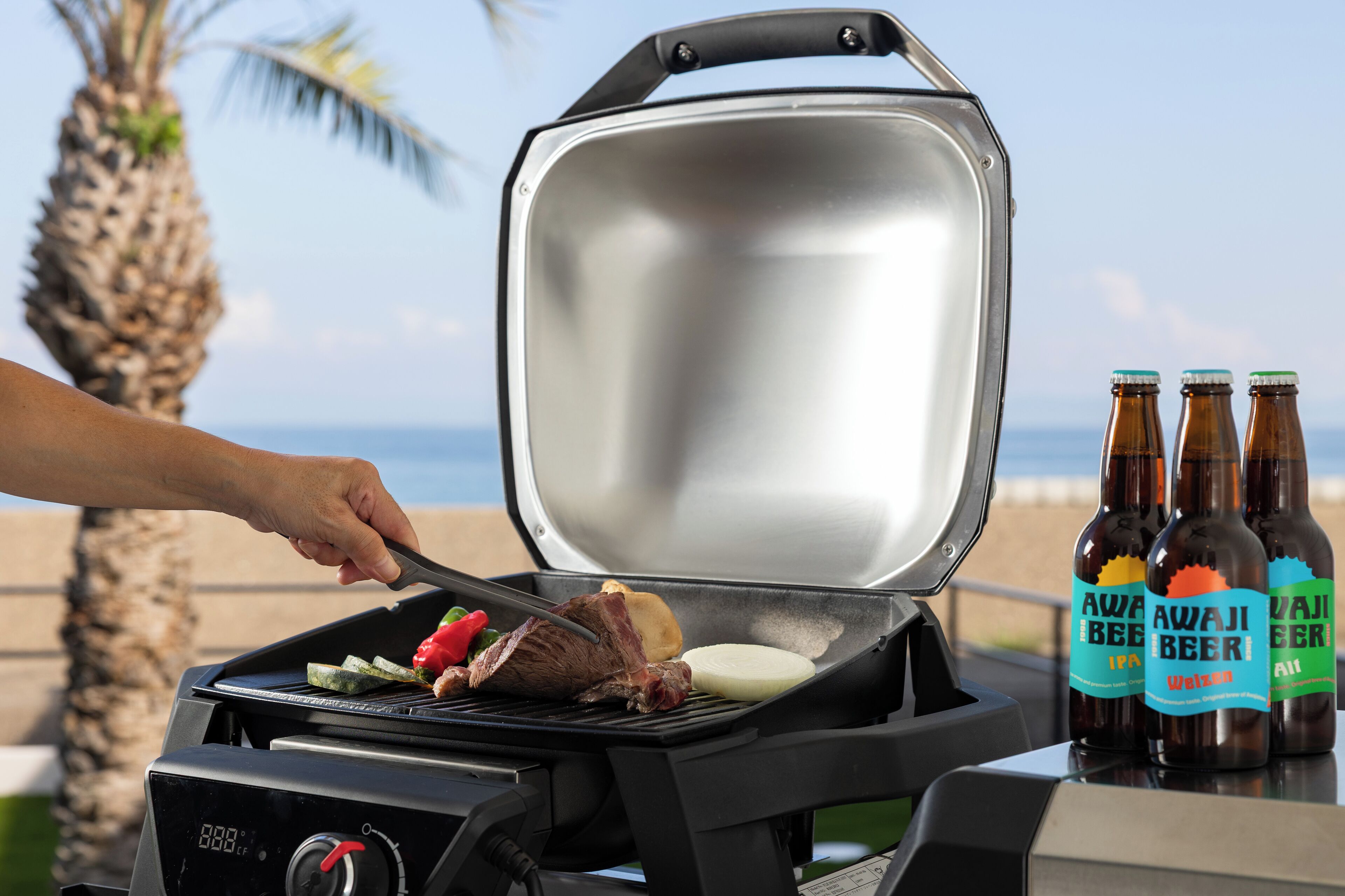 Ein tragbarer Grill mit Fleisch und Gemüse, das darauf zubereitet wird, und drei Flaschen Bier mit dem Aufdruck 'Awa Jibe Beer' auf einem Tisch.