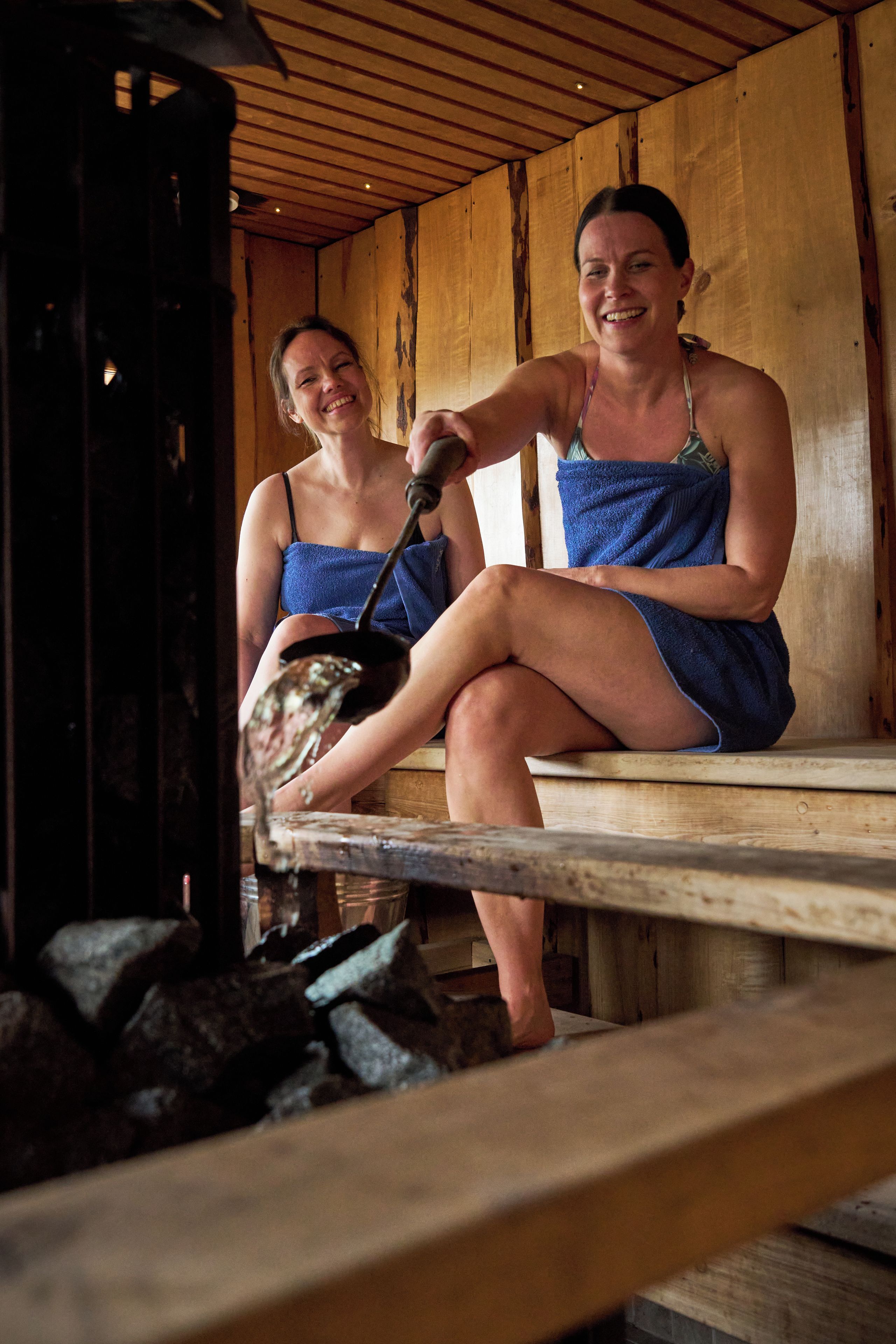 Two individuals seated on wooden benches in a rustic cabin with a wood-burning stove in the foreground.