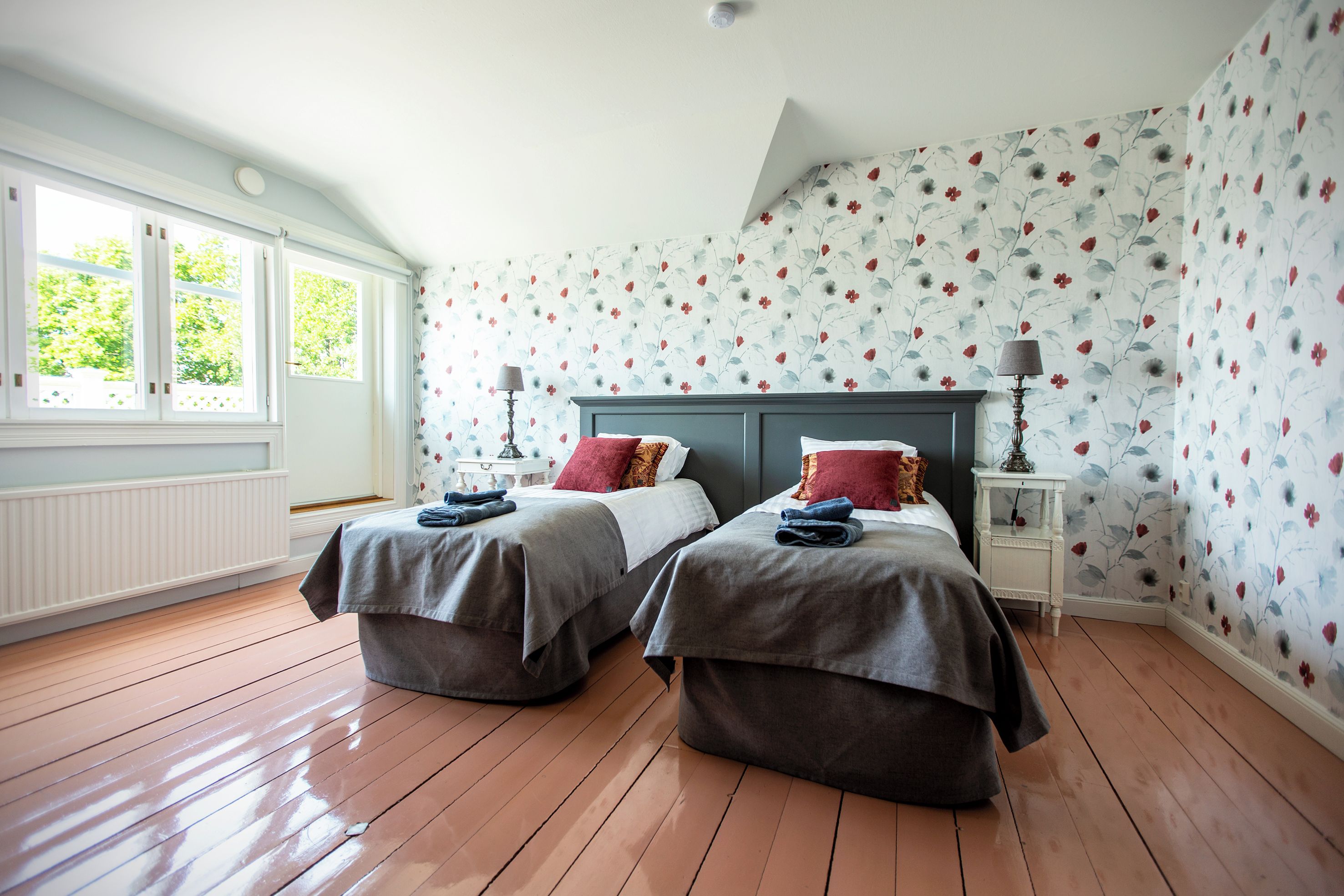A bedroom with two beds, a window with a view, and floral wallpaper.
