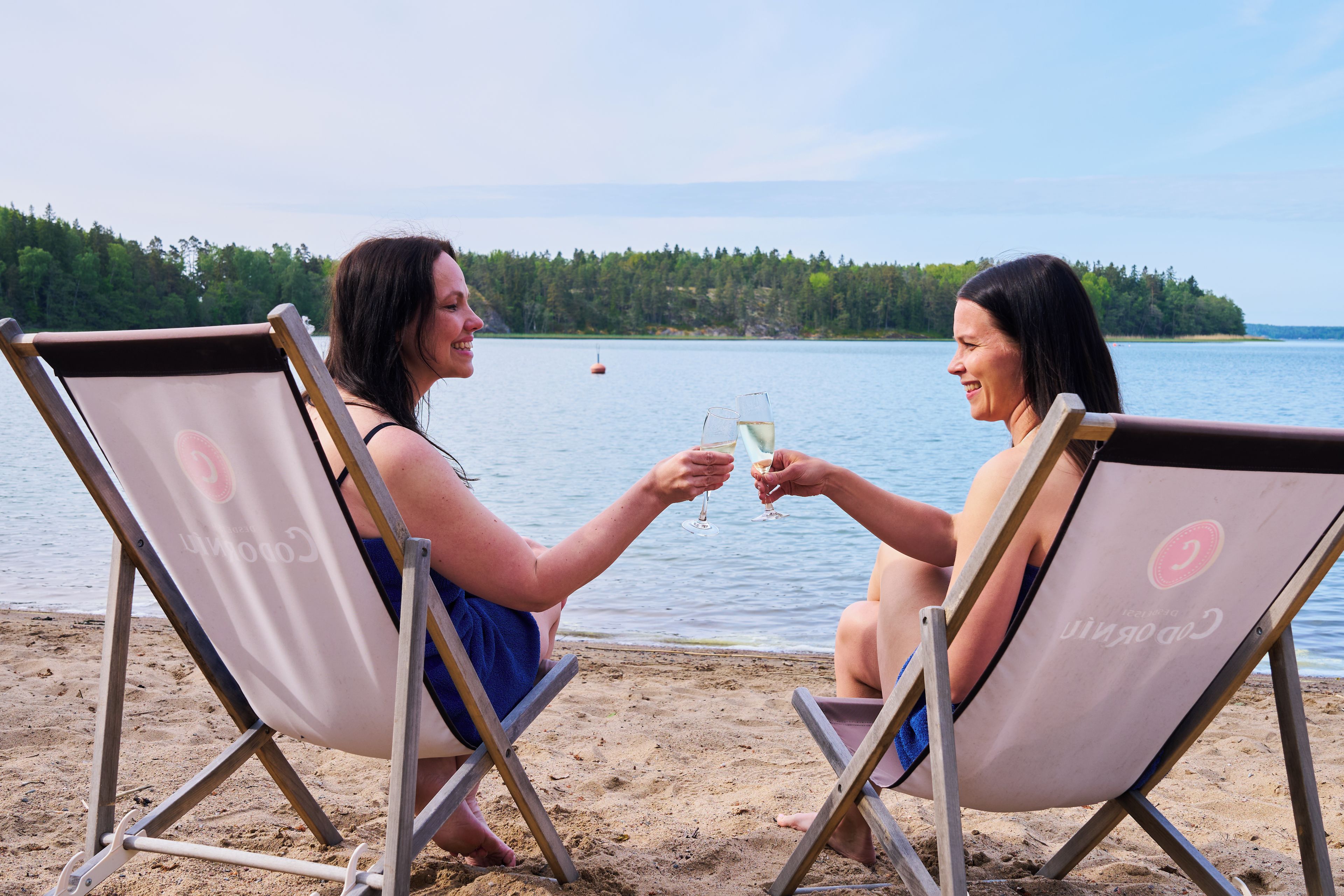 Two individuals seated on beach chairs facing a body of water with a forested island in the background.
