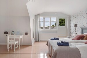 Un dormitorio con cama, escritorio y silla. Se ve una ventana con cortinas, un radiador y una lámpara sobre una mesita de noche.