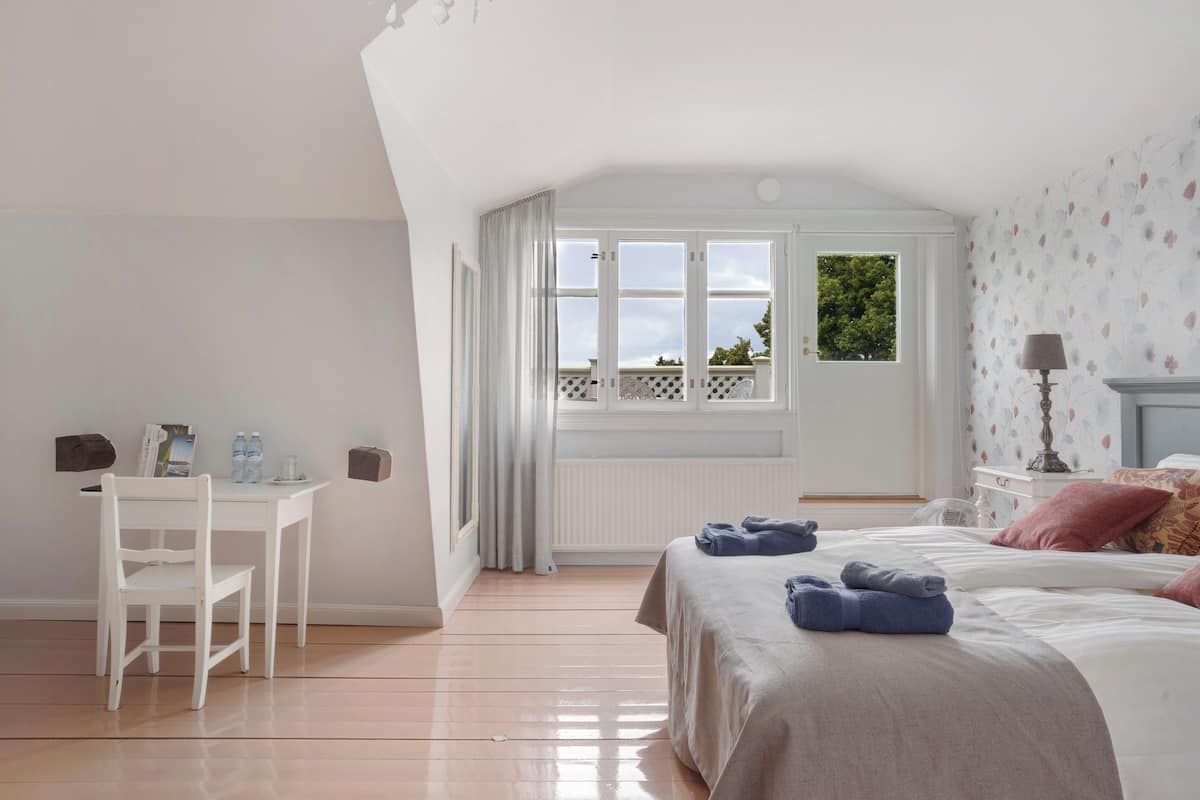 A bedroom with a bed, a desk, and a chair. There is a window with curtains, a radiator, and a lamp on a bedside table.
