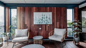 Ein modernes Interieur mit Holzwandverkleidung, zwei Korbstühlen, einem kleinen runden Tisch und großen Fenstern mit Blick auf Grünflächen.