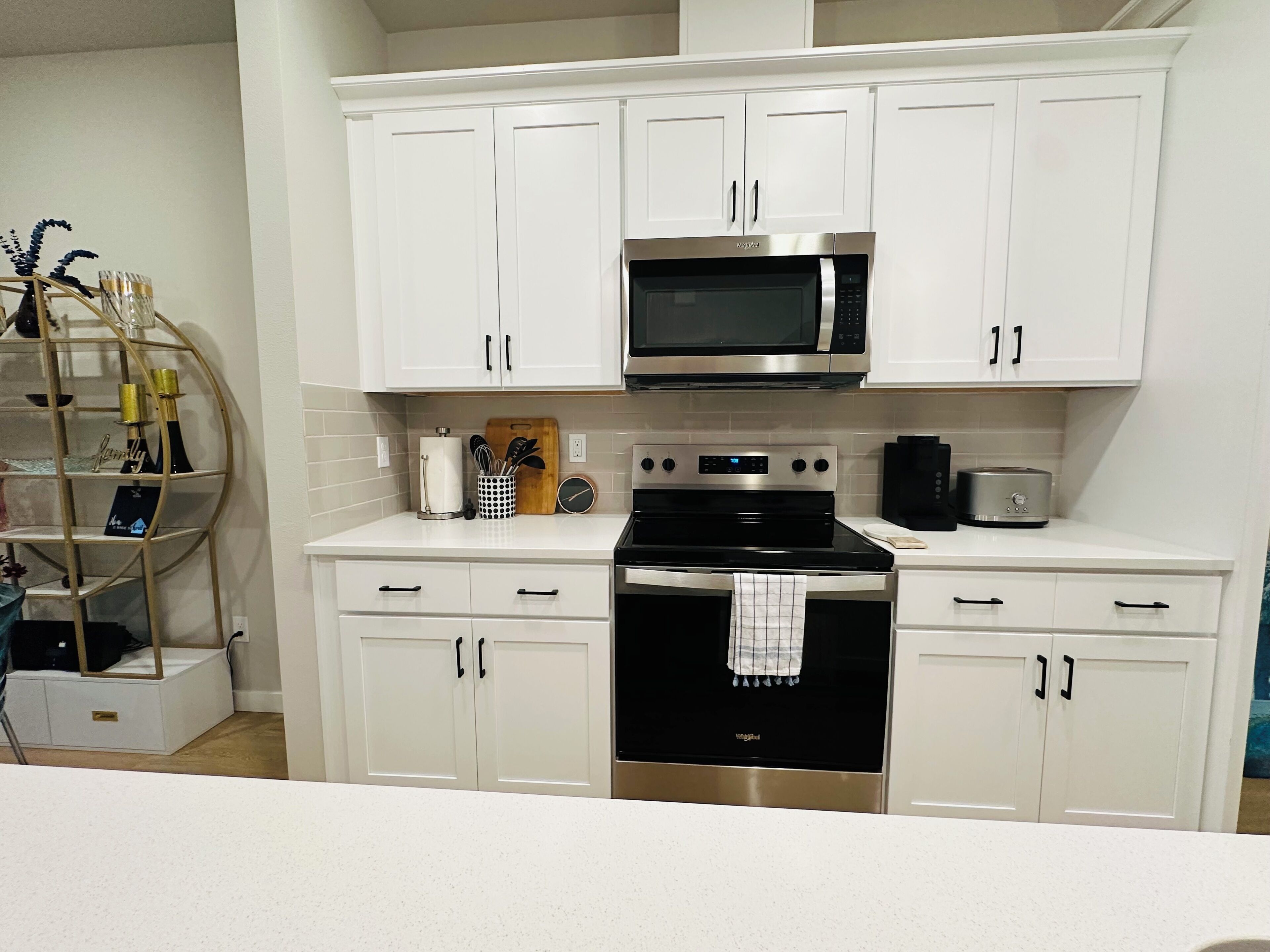 A modern kitchen with white cabinetry, a stainless steel microwave, and an oven. There is a wooden cutting board with utensils on the countertop.