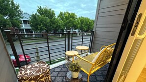 A balcony with a yellow metal chair and a patterned ottoman, a small table with a potted plant, and a view of a street with houses and a red car. - Charming townhome with AC in Hillsboro (Hillsboro)