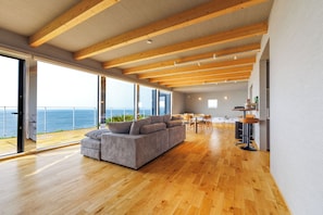 A spacious living area with a large sofa, wooden flooring, and a view of the sea through large windows. - Yunagi  (Ishikari)