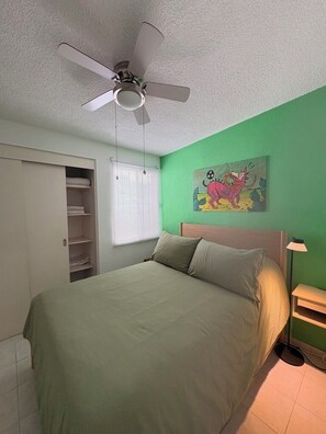 A bedroom with a bed, a ceiling fan, a closet, a window with blinds, and a wall painting.