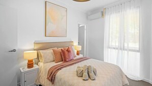 A neatly arranged bedroom with a bed, bedside lamps, a large window with sheer curtains, and a wall-mounted air conditioning unit. - Best Location Armadale Sleeps 6 (Perth)