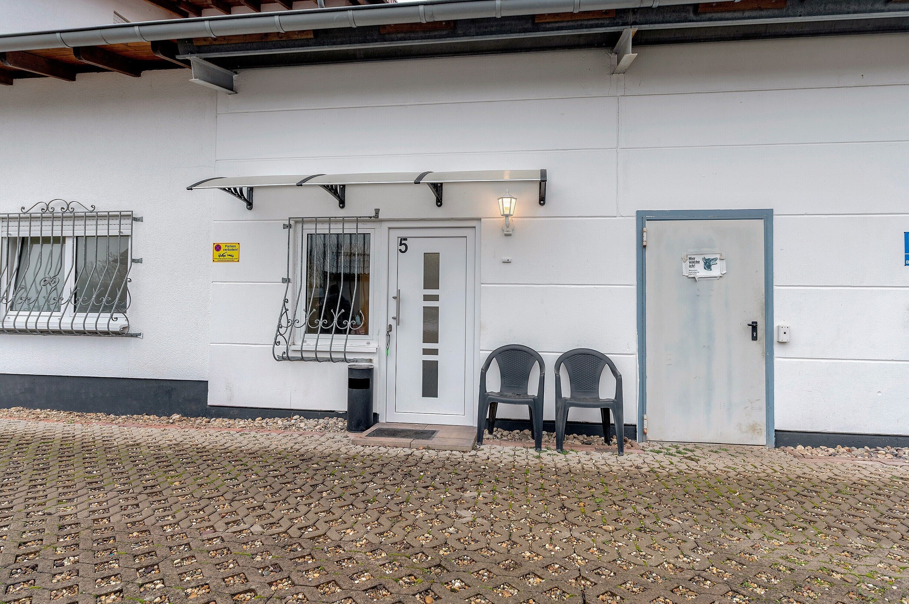 Un edificio con fachada blanca, una puerta numerada, dos sillas afuera y un bote de basura pequeño.