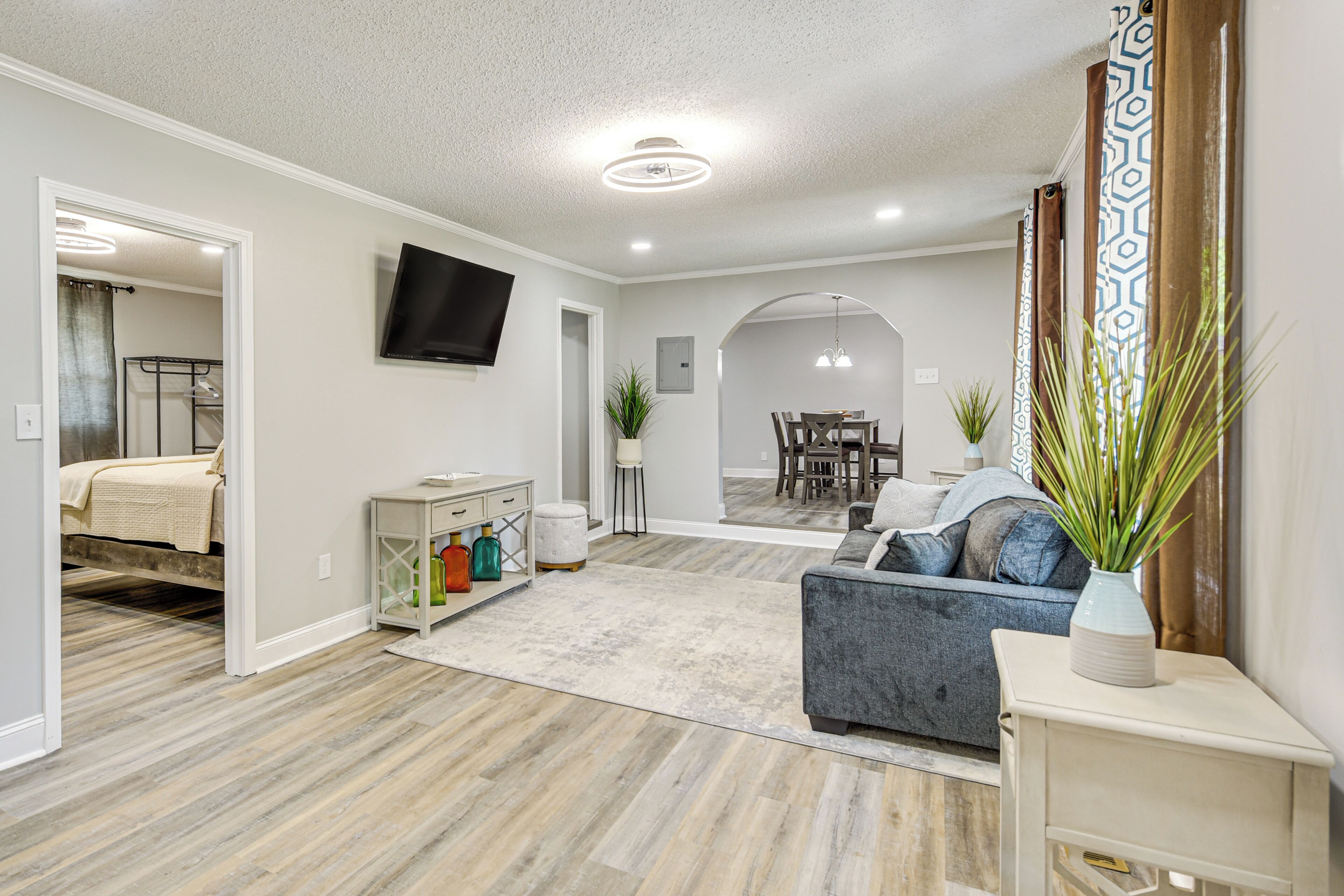 A spacious living area with a grey sofa, a TV mounted on the wall, and a dining area in the background.