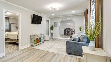 A spacious living area with a grey sofa, a TV mounted on the wall, and a dining area in the background.