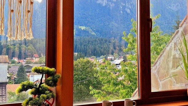 Una habitación acogedora con un amplio ventanal que da a un paisaje montañoso, un bonsái y un otoman tejido.