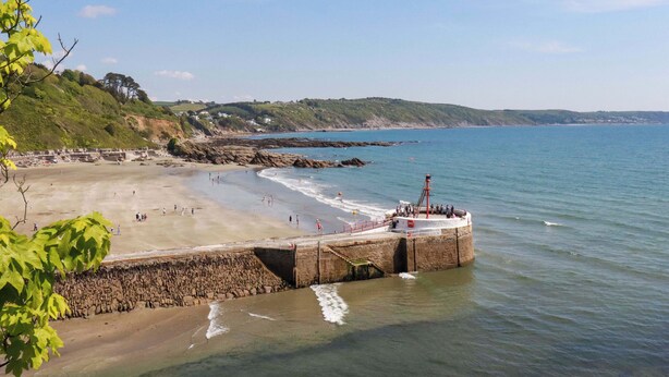 A coastal beach with a stone pier extending into the sea, surrounded by hills and houses in the background. - Woodview (Looe)