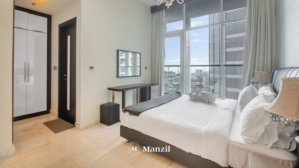 A modern hotel room with a large bed, a desk, and a view of the city through large windows. - Manzil - Luxury 2BR w Pool and Burj View (Dubai)