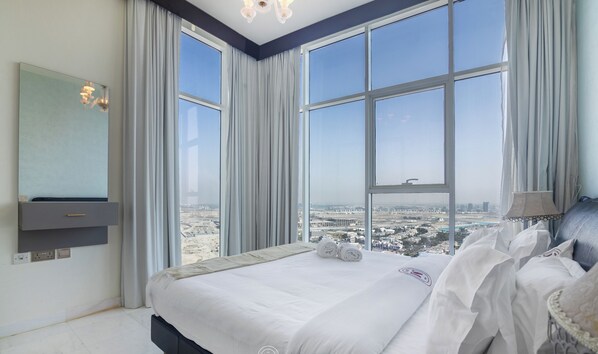 Un dormitorio con una cama grande, un televisor montado en la pared y vistas de un paisaje urbano a través de amplios ventanales.