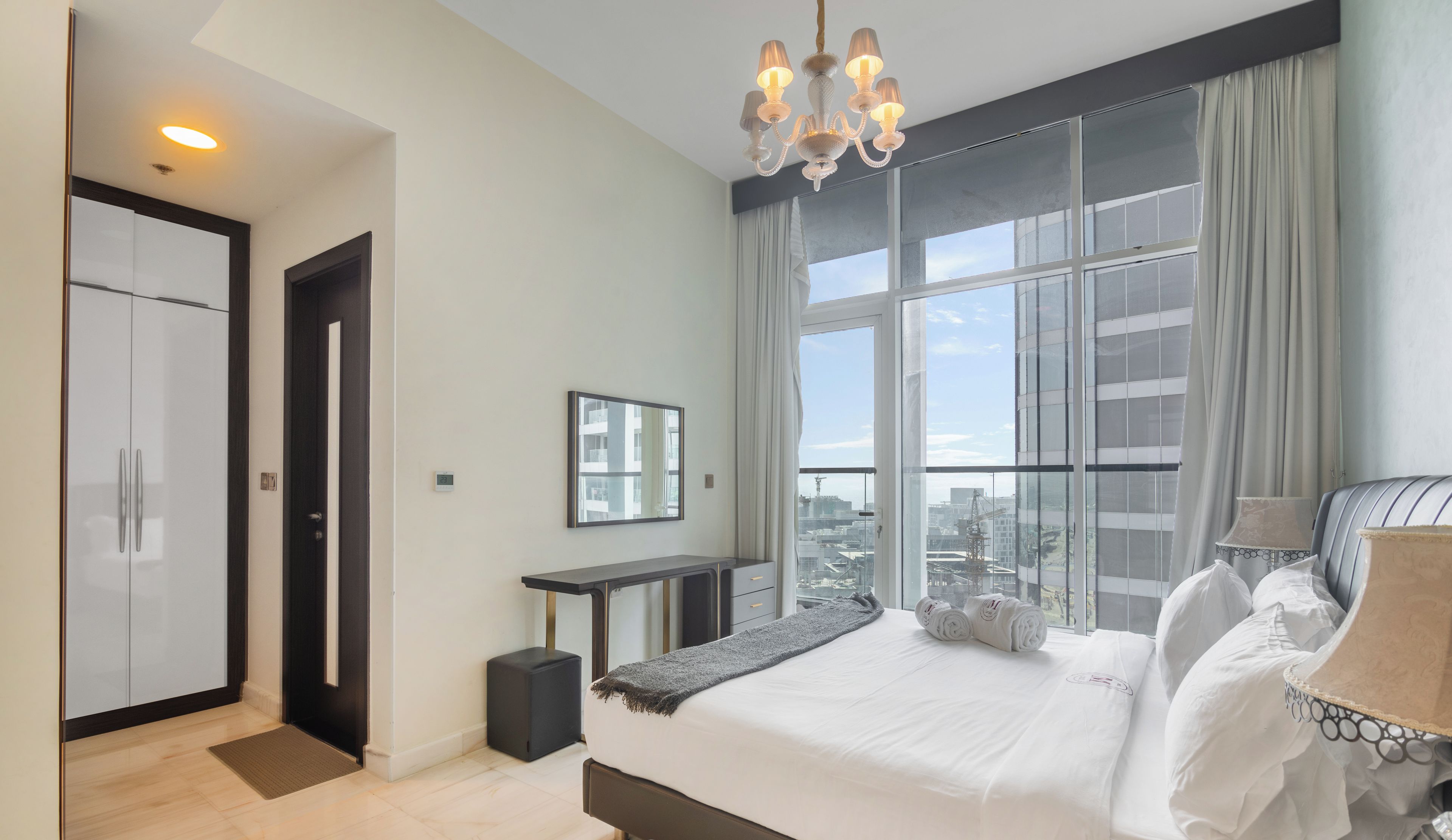 A modern hotel room with a large bed, a desk, and a view of the city through large windows.