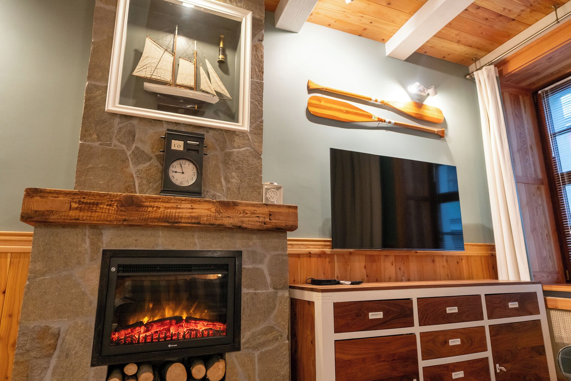 A stone fireplace with logs, a clock, a framed ship model, and a mounted paddleboard.