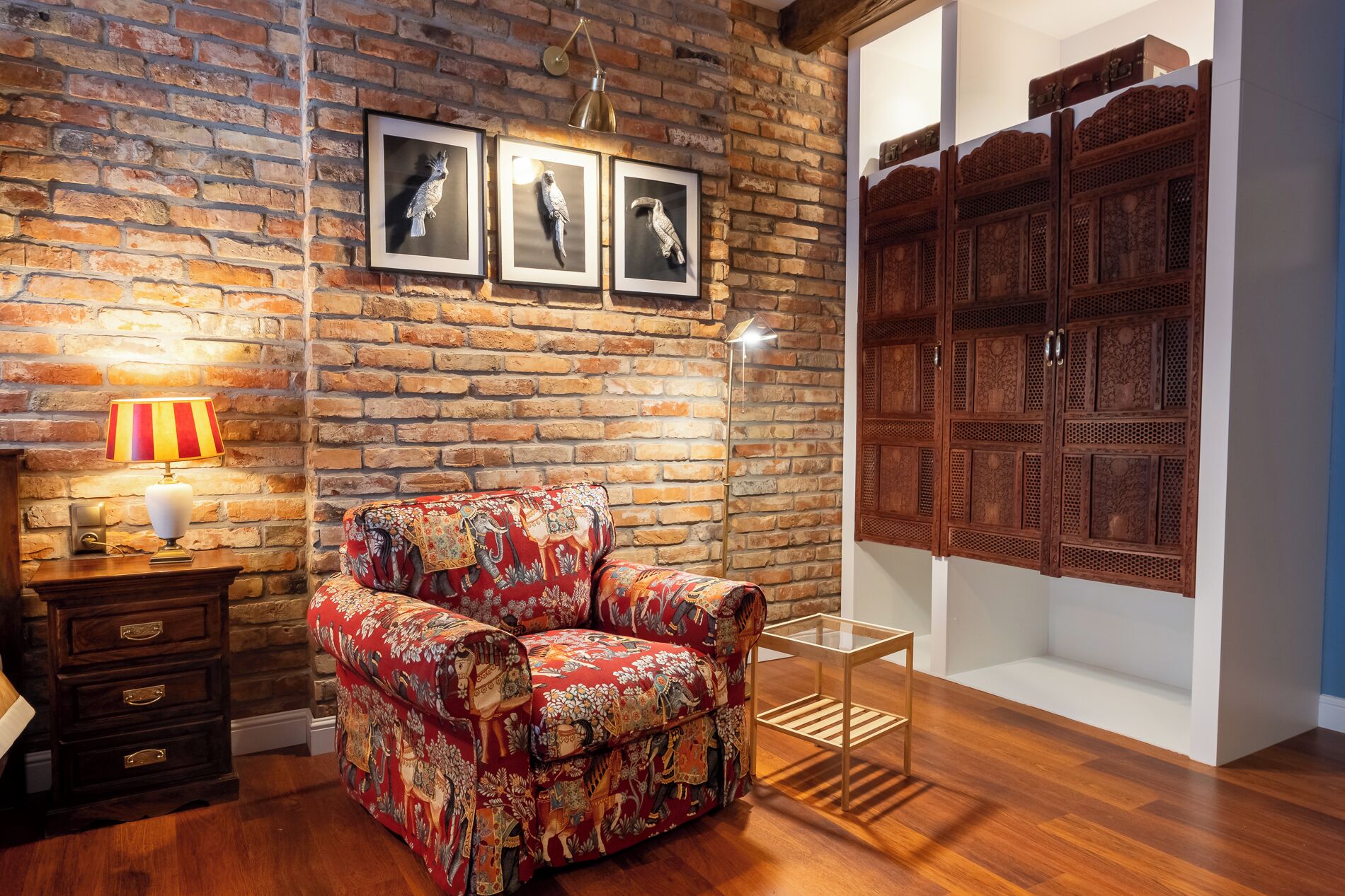 Une pièce avec un mur en briques, un fauteuil à motifs, un meuble en bois et des œuvres d’art encadrées.